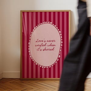 Könnte beinhalten: Gerahmter Druck mit einem rosa-roten Streifenhintergrund. Der Druck zeigt eine ovale Form mit dem Zitat "Love's never wasted when it's shared" in einer Schrift. Der Rahmen ist hellbraun.