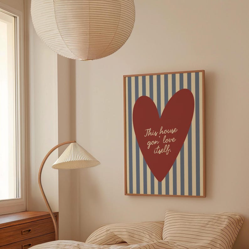 May include: A framed wall art print featuring a large red heart on a background of blue and beige vertical stripes. The text "This house gon' love itself" is written inside the heart. The artwork is in a room with a lamp and a bed.