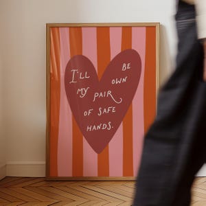 May include: Framed art print in a wooden frame. The artwork features a large maroon heart with the words "I'll be my own pair of safe hands" in white. The background has vertical stripes of pink and orange.