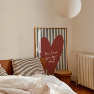 May include: A bedroom scene featuring a framed poster with a red heart and the text "This house gon' love itself." The poster has a blue and white striped background. A striped pillow and a round wooden stool are also visible.