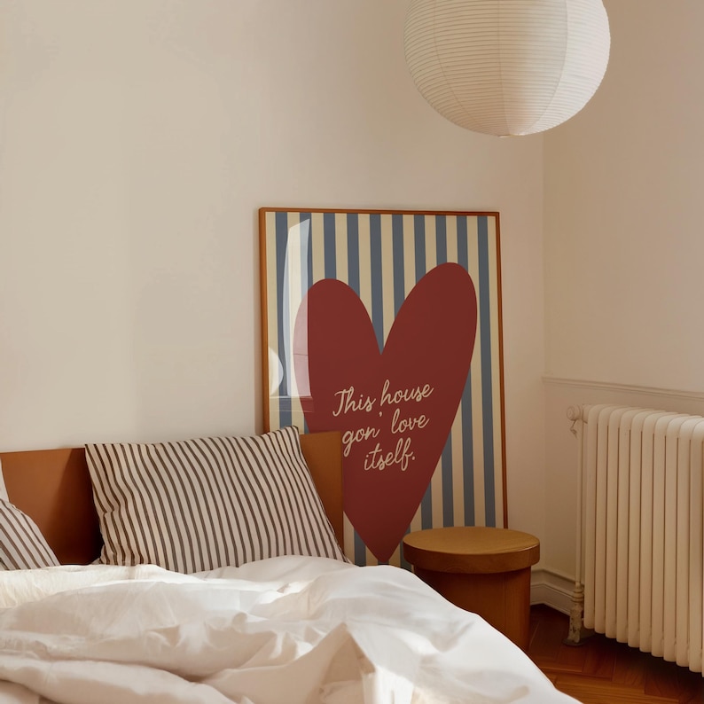 May include: A bedroom scene featuring a framed poster with a red heart and the text "This house gon' love itself." The poster has a blue and white striped background. A striped pillow and a round wooden stool are also visible.