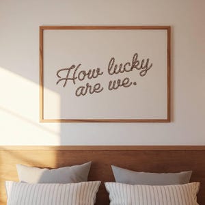 May include: A framed print with the words "How lucky are we" written in a rope-style font. The artwork has a light background and a wooden frame. The print is displayed above a bed with pillows.