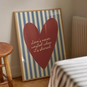 May include: A framed print featuring a large red heart on a striped background of blue and cream. The heart contains the quote, "Love's never wasted when it's shared." The frame is a light brown color. The print is leaning against a wall.
