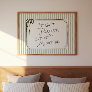 May include: A framed print with the text "It isn't perfect, but it might be." The print has a green and white striped background and a wooden frame. A green ribbon is tied to the upper left corner of the frame. The print is hanging above a bed.