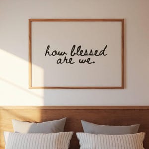 Könnte beinhalten: Ein gerahmter Druck mit dem Schriftzug "how blessed are we" in schwarzer Schrift. Das Kunstwerk hat einen hellen Hintergrund und einen Naturholzrahmen. Der Druck wird über einem Bett mit Kissen angezeigt.