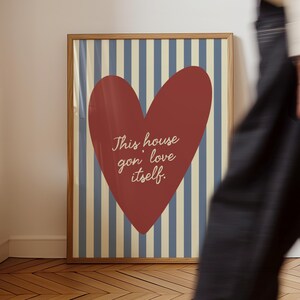 May include: A framed art print featuring a large red heart with the text "This house gon' love itself" on a background of blue and cream vertical stripes. The frame is made of wood.