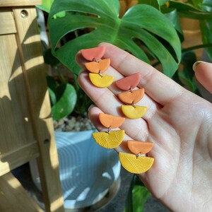 Puede incluir: Un par de pendientes hechos a mano de arcilla polimérica con un diseño en capas. Los pendientes están hechos de tres semicírculos en tonos naranja, amarillo y un amarillo texturizado.
