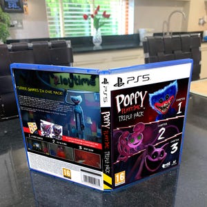 May include: A PS5 game case for "Poppy Playtime Triple Pack" is open, revealing the game's cover art. The cover features a blue monster with sharp teeth and the text "Poppy Playtime Triple Pack" with chapters 1, 2, and 3. The case is blue.