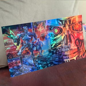 May include: A holographic print featuring the Terminator T-800 robot holding a weapon, with a skull graphic on the right side. The artwork has a vibrant color scheme of blues, reds, and yellows, with the text "TERMINATOR 2 JUDGMENT DAY".