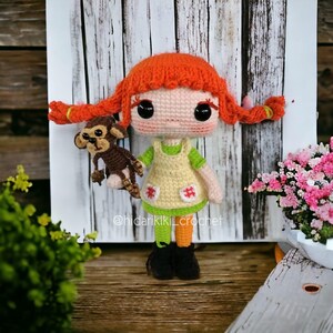 May include: A handmade crochet doll with red pigtails, wearing a yellow dress with green accents, and holding a brown monkey. The doll has large black eyes and is set against a wooden backdrop. The text "@hidarikiki_crochet" is visible.