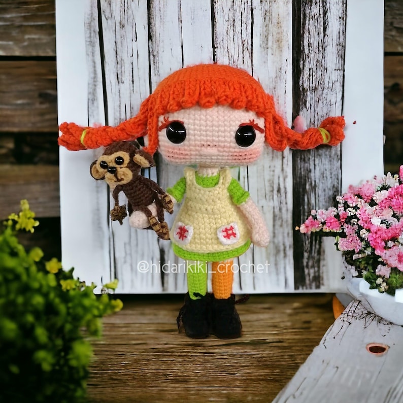 May include: A handmade crochet doll with red pigtails, wearing a yellow dress with green accents, and holding a brown monkey. The doll has large black eyes and is set against a wooden backdrop. The text "@hidarikiki_crochet" is visible.