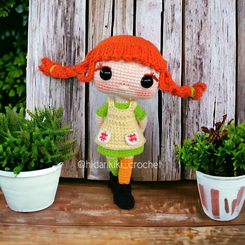 May include: A handmade crochet doll with orange pigtails, wearing a yellow dress with red hashtag accents, green shirt, orange socks, and black boots. The doll has large black eyes and is set against a wooden background. The text "@hidarikiki_crochet" is visible.
