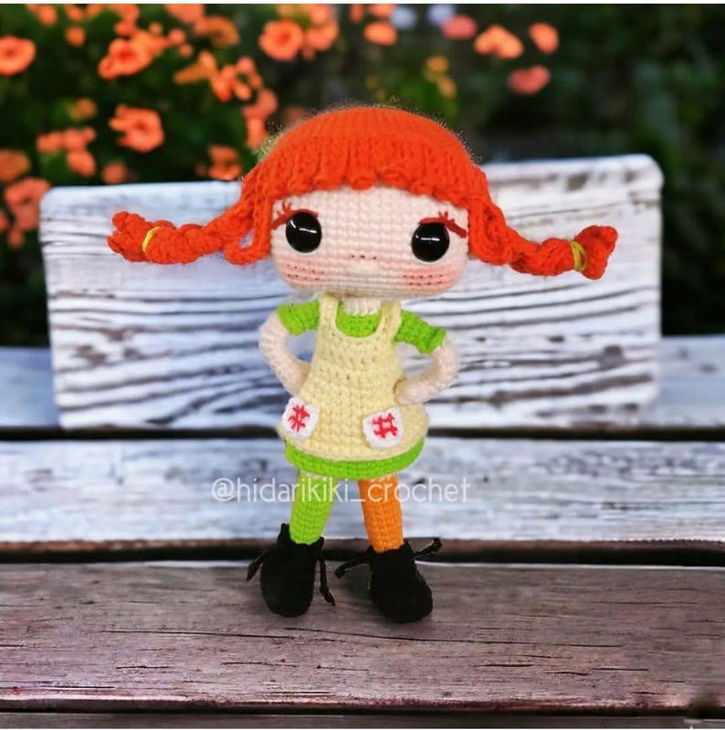 May include: A handmade crochet doll with bright red pigtails, large black eyes, and a light skin tone. The doll wears a yellow dress with green accents, orange and green striped legs, and black shoes. The doll is standing on a wooden surface.