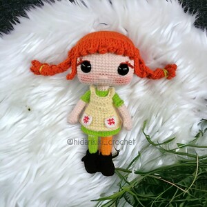 May include: A handmade crochet doll with bright orange hair styled in pigtails. The doll wears a green shirt, a yellow dress with red hashtag accents, orange and green leggings, and black boots. The doll is set against a white, fluffy background.