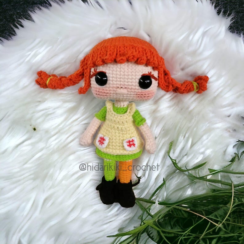 May include: A handmade crochet doll with bright orange hair styled in pigtails. The doll wears a green shirt, a yellow dress with red hashtag accents, orange and green leggings, and black boots. The doll is set against a white, fluffy background.