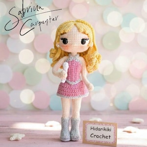 May include: A handmade crochet doll with blonde curly hair, wearing a pink dress and gray boots, holding a microphone. The doll has black eyes and is standing on a pink surface. The text "Sabrina Carpenter" is written on the top left.