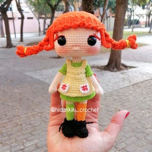 May include: A handmade crochet doll with bright orange pigtails, large black eyes, and a green and yellow dress. The doll has black shoes and orange and green striped legs. The doll is held in a hand. The doll is a Pippi Longstocking inspired design.