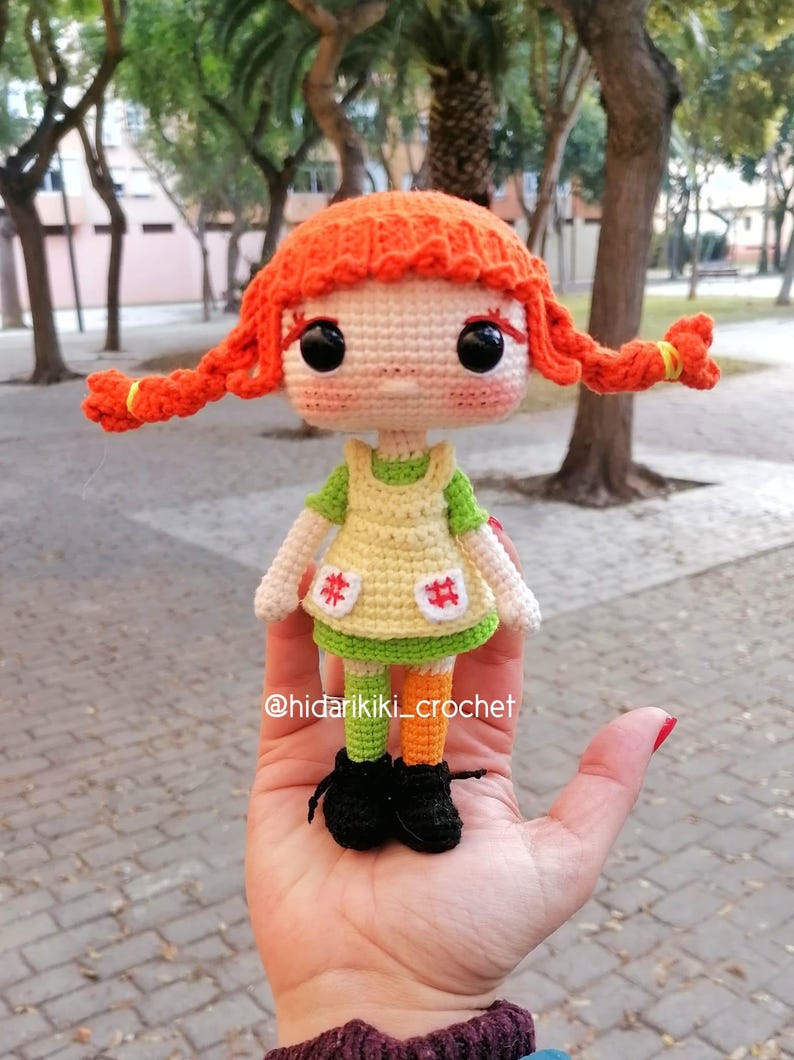 May include: A handmade crochet doll with bright orange pigtails, large black eyes, and a green and yellow dress. The doll has black shoes and orange and green striped legs. The doll is held in a hand. The doll is a Pippi Longstocking inspired design.