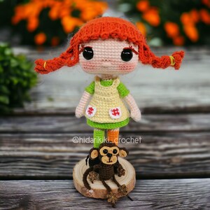 May include: A handmade crochet doll with red braided hair, wearing a yellow dress with green sleeves and a green skirt. A small brown crochet monkey sits on a wooden base in front of the doll. The doll has large black eyes and a sweet expression.