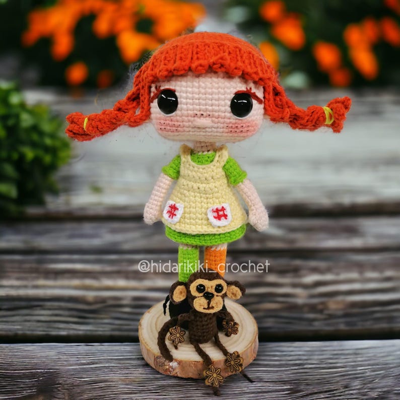 May include: A handmade crochet doll with red braided hair, wearing a yellow dress with green sleeves and a green skirt. A small brown crochet monkey sits on a wooden base in front of the doll. The doll has large black eyes and a sweet expression.