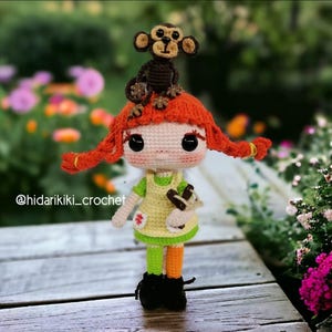 May include: A handmade crochet doll with red pigtails and a small monkey on its head. The doll wears a yellow dress, green and orange striped leggings, and black boots. The doll is holding a small brown object. The text "@hidarikiki_crochet" is visible.