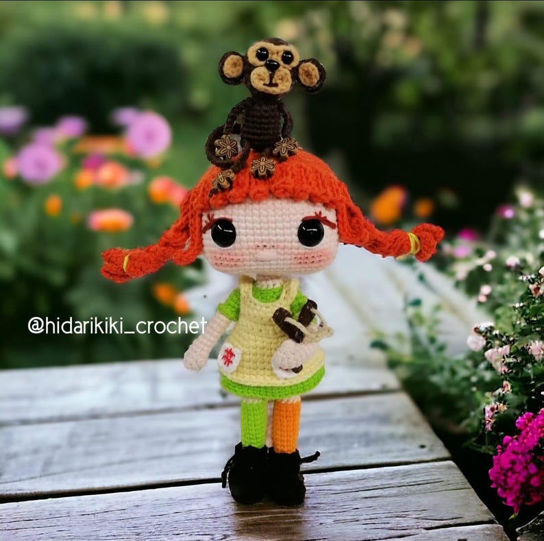 May include: A handmade crochet doll with red pigtails and a small monkey on its head. The doll wears a yellow dress, green and orange striped leggings, and black boots. The doll is holding a small brown object. The text "@hidarikiki_crochet" is visible.