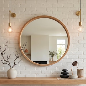 May include: A large round mirror with a light wood frame mounted on a white brick wall above a floating wooden shelf. The shelf holds a white vase with bare branches, a stack of three black stones, a small ribbed bowl, and a textured beige pot.