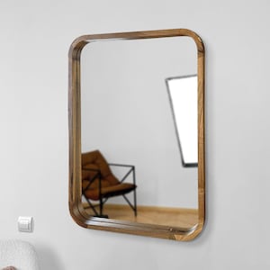 May include: A rectangular wooden framed mirror with a rounded edge. The mirror is hanging on a white wall.