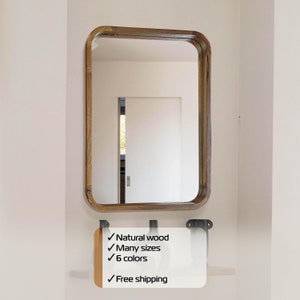 May include: A rectangular wooden framed mirror with rounded edges. The mirror is hanging on a white wall. The frame is a dark brown wood. The mirror reflects a white door with a window in the background.