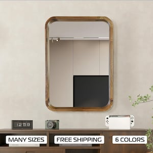 Peut inclure: Un miroir rectangulaire encadré de bois avec un dessus et un bas arrondis. Le miroir est suspendu à un mur beige clair. Le miroir reflète une pièce avec une télévision noire et des lumières de plafond blanches. Sous le miroir se trouve une étagère en bois avec une horloge numérique, un appareil photo et une console Nintendo Switch blanche.