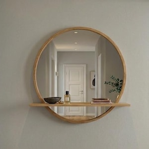 Peut inclure: Un miroir rond encadré de bois avec une petite étagère, fixé sur un mur gris clair. L'étagère contient un petit bol, un flacon de parfum et des livres. Le miroir reflète un couloir avec un sol en bois et un tapis aux tons neutres.