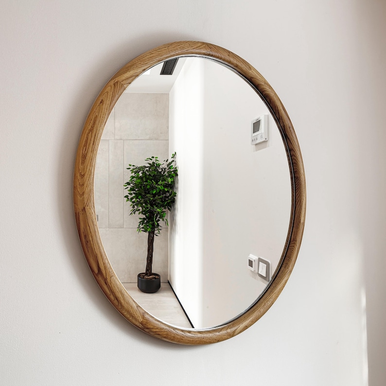 May include: A round wooden framed mirror with a natural wood finish. The mirror reflects a room with a potted plant and white walls.