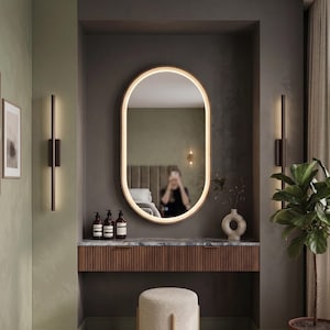 Può includere: Uno specchio da parete ovale con illuminazione LED perimetrale integrata, posizionato sopra una console sospesa in legno con piano in marmo, caratterizzato da listelli verticali sul cassetto e due sottili applique da parete in metallo scuro.