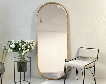 May include: A large, oval-shaped full-length mirror with a light wood frame. The mirror is positioned against a light gray wall. A black metal chair with a cream-colored seat and back is next to the mirror. A black metal plant stand with white flowers is also in the image.