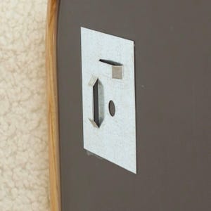 May include: A rectangular metal wall mounting bracket attached to a dark gray surface with a light wood-toned frame edge. The bracket features a vertical slot, a horizontal slot, and a small circular hole for secure installation on a wall.