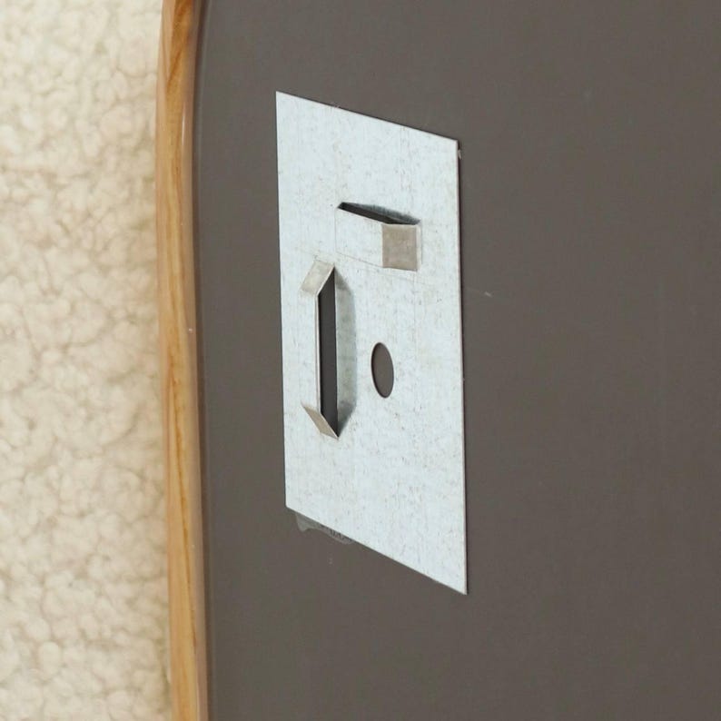 May include: A rectangular metal wall mounting bracket attached to a dark gray surface with a light wood-toned frame edge. The bracket features a vertical slot, a horizontal slot, and a small circular hole for secure installation on a wall.