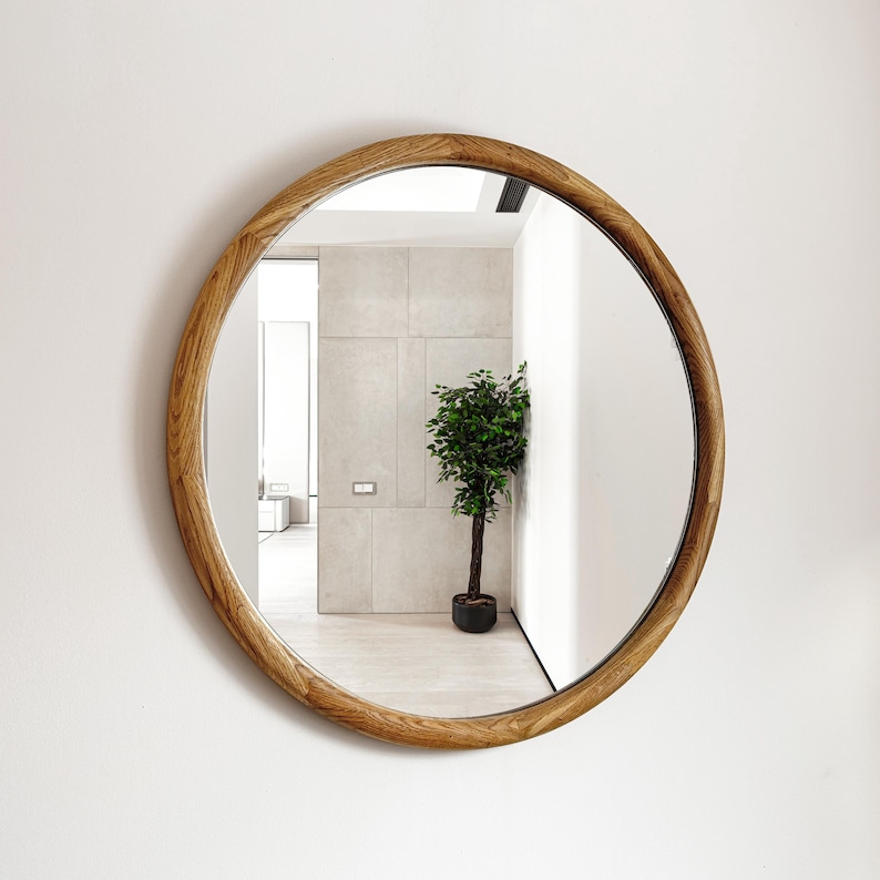 May include: A round wooden framed mirror with a natural wood finish. The mirror reflects a room with a potted plant and a doorway.