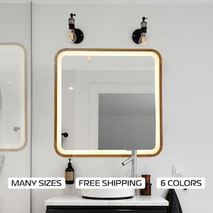 May include: A modern bathroom vanity with a rectangular mirror with a wooden frame. The mirror has a white LED light around the perimeter. The vanity has a white sink and a chrome faucet. There are two black wall sconces with Edison bulbs on either side of the mirror. The text "MANY SIZES", "FREE SHIPPING", and "6 COLORS" is visible at the bottom of the image.