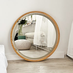 May include: A large circular wall mirror featuring a smooth, light-toned wooden frame. The reflective glass surface shows a clear view of a room interior, including a white knitted pouf, a glass vase with white flowers, and a white radiator.