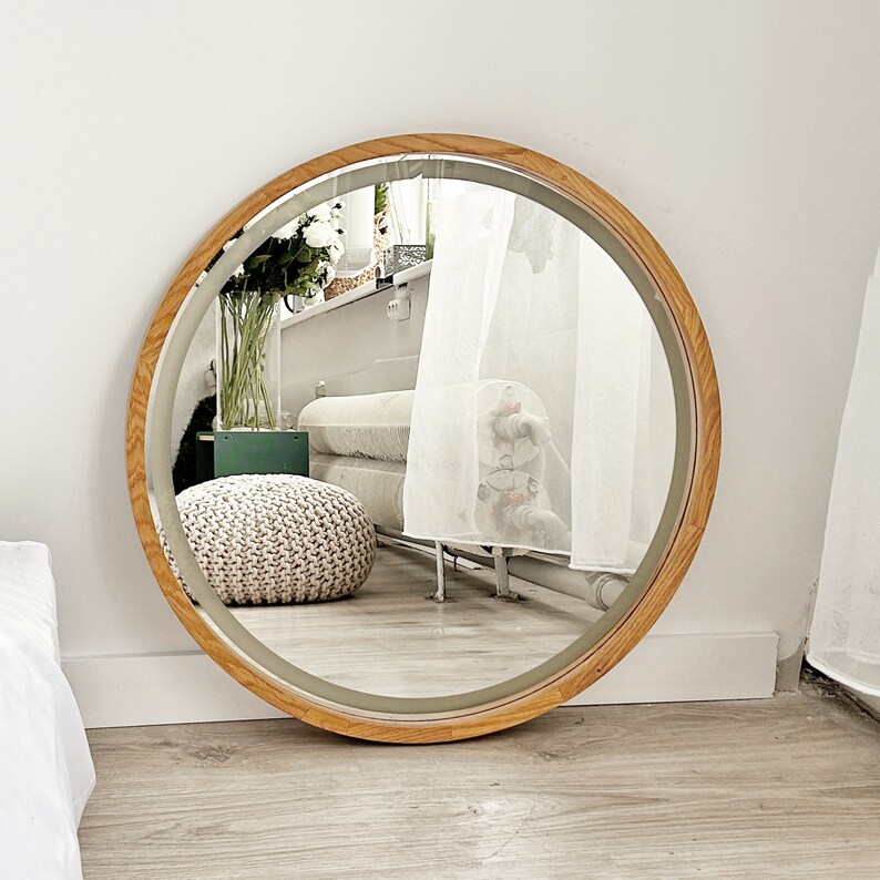 May include: A large circular wall mirror featuring a smooth, light-toned wooden frame. The reflective glass surface shows a clear view of a room interior, including a white knitted pouf, a glass vase with white flowers, and a white radiator.