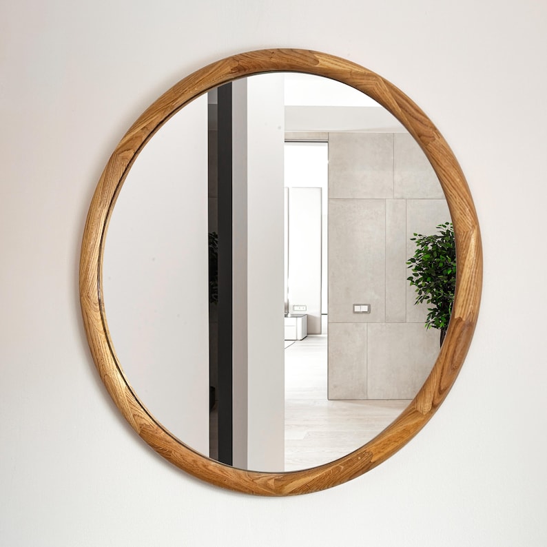 May include: A round mirror with a natural wood frame. The mirror reflects a hallway with a door and a plant.