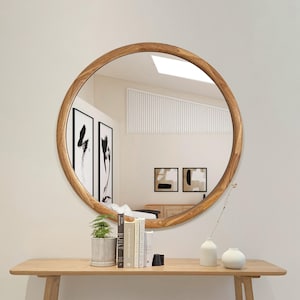 May include: A round wooden framed mirror with a natural wood finish. The mirror is hanging on a white wall above a wooden console table with a vase and books.