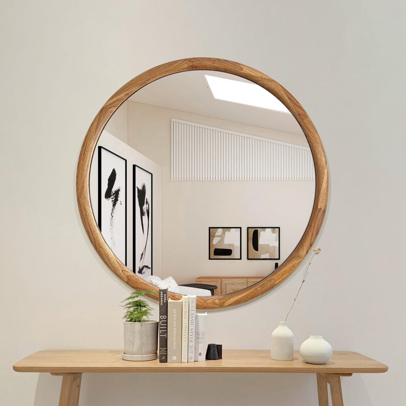 May include: A round wooden framed mirror with a natural wood finish. The mirror is hanging on a white wall above a wooden console table with a vase and books.