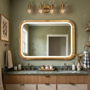 May include: A rectangular wall mirror with rounded corners and a wooden frame featuring an integrated LED light strip. The mirror is mounted above a double vanity with a green countertop, two brass faucets, and a light wood fluted cabinet base.