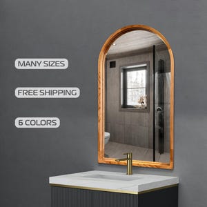 Op de afbeelding: Gebogen spiegel met een lichtbruine houten lijst. De spiegel reflecteert een badkamer met een raam en wastafel. De teksten "MANY SIZES" en "6 COLORS" zijn zichtbaar.