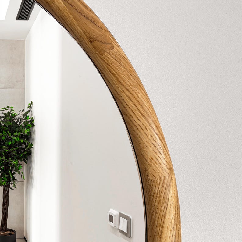 May include: A round mirror with a natural wood frame. The frame is thick and has a rounded edge. The mirror is reflecting a white wall with a light switch.