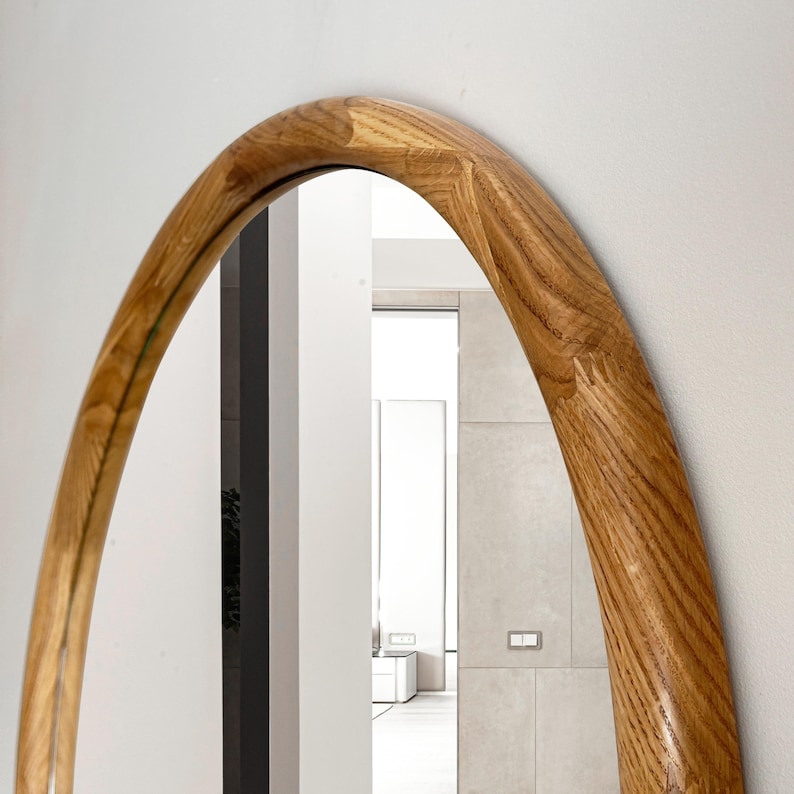 May include: A large oval mirror with a thick, natural wood frame. The frame is a warm brown colour and has a slightly curved shape. The mirror reflects a white room with a door and a window.