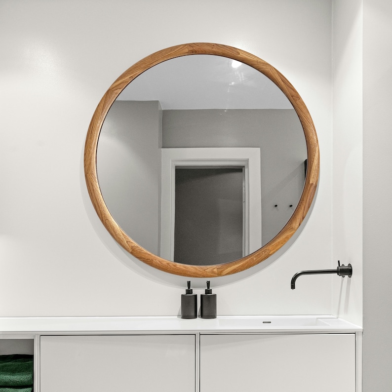 May include: A round wooden framed mirror with a white wall and a white countertop with two black soap dispensers and a black faucet.