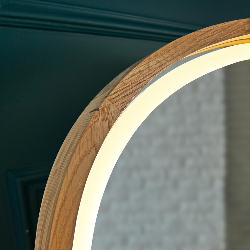 May include: A circular mirror featuring a light-toned wooden frame with a visible grain pattern. An integrated LED light strip runs along the inner edge of the frame, emitting a warm, bright glow that illuminates the reflective surface of the mirror.