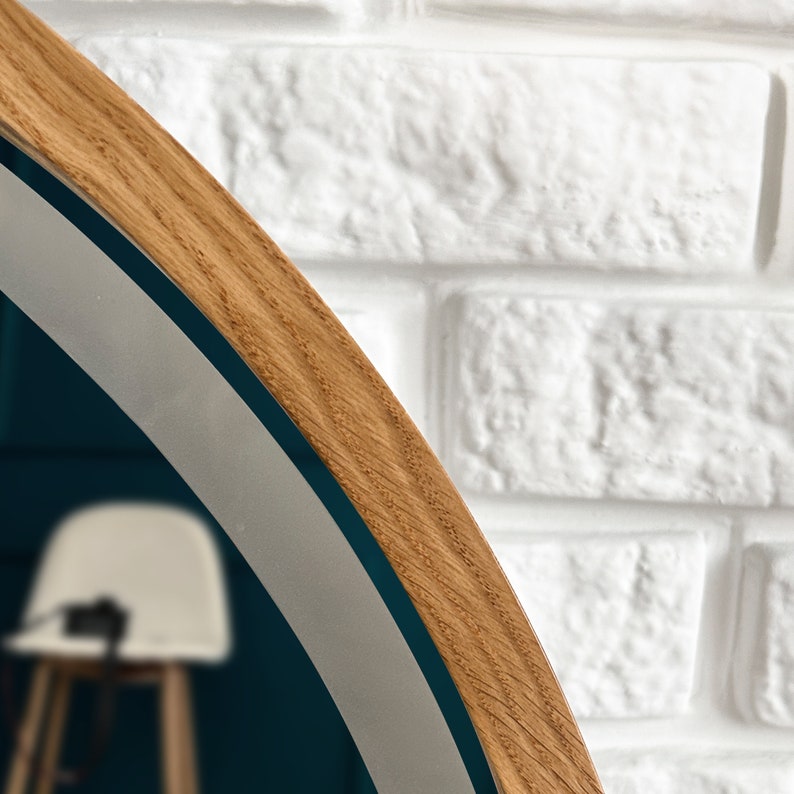 May include: A circular mirror featuring a light brown wood frame with a visible grain pattern. The mirror surface reflects a white chair and a dark blue wall, while the outer edge of the frame is positioned against a white textured brick wall.
